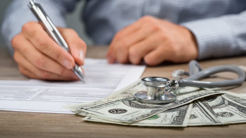 Person filling out insurance forms with stethoscope and cash on the table, symbolizing healthcare costs