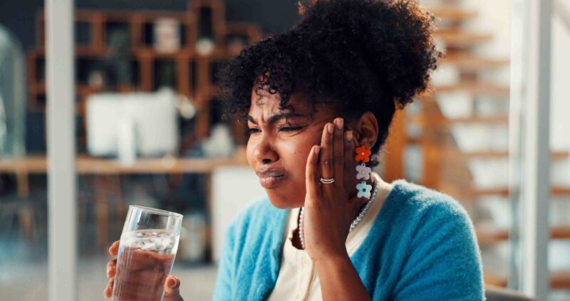 Person holding their cheek with facial swelling discomfort while holding a glass of water
