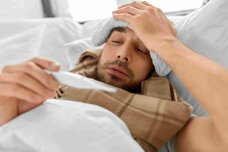 Person lying in bed with a fever, checking a thermometer while feeling unwell